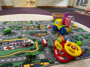 An image of a playmat depicting a city, full of roads, with various car and truck toys on it, and a little toy steering wheel.